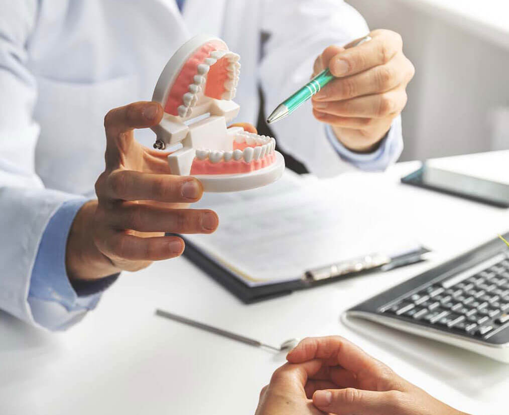 Dentist in white coat demonstrates dental model and instrument to patient at desk with laptop.
