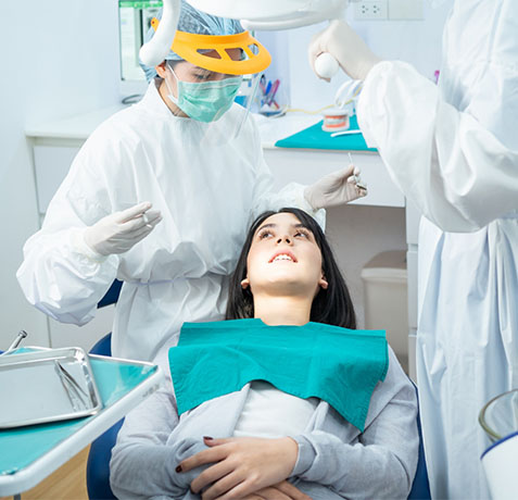 Dental professional examining patient in clinical setting with protective equipment and instruments.